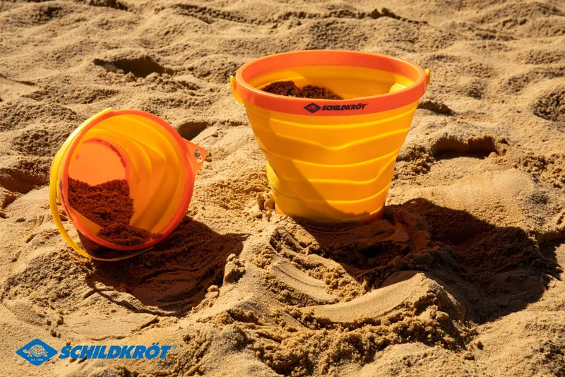 Seau de plage rétractable orange Schildkröt – mise en situation : deux seaux orange sur la plage, l’un couché, l’autre debout