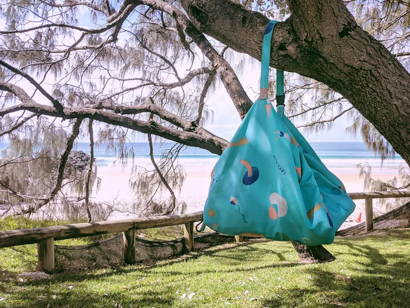 Tapis de jeu et sac de rangement d’extérieur Play and Go – motif ballons – vue du sac en version rangement accroché à un arbre, avec une plage en arrière-plan