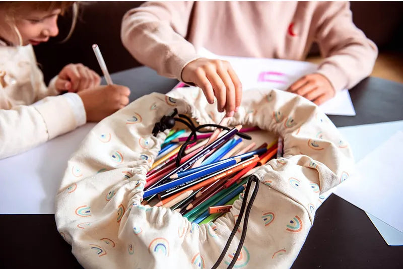 Mini sac de rangement et tapis de jeux motif arc-en-ciel de Playandgo - vue du sac ouvert avec des crayons et des enfants qui dessinent en arrière plan