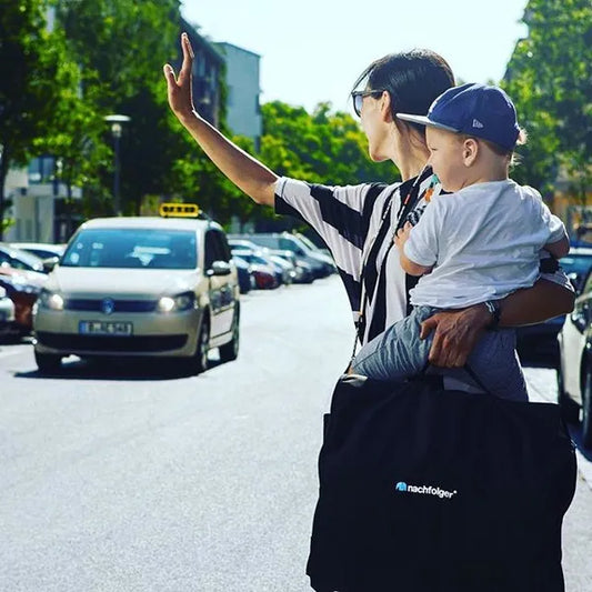 Siège auto gonflable HyFive One TT CITY de Nachfolger – vue en situation : une dame appelle un taxi, son bébé dans les bras et le siège rangé dans son sac.