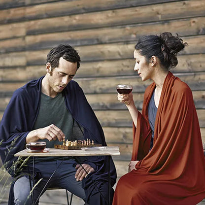 Couverture de voyage en laine et soie – mise en situation : la couverture bleu marine est portée par un homme assis, la terracotta par une femme assise jouant aux échecs.