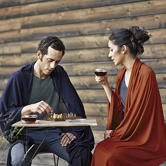 Couverture de voyage en laine et soie – mise en situation : la couverture bleu marine est portée par un homme assis, la terracotta par une femme assise jouant aux échecs.